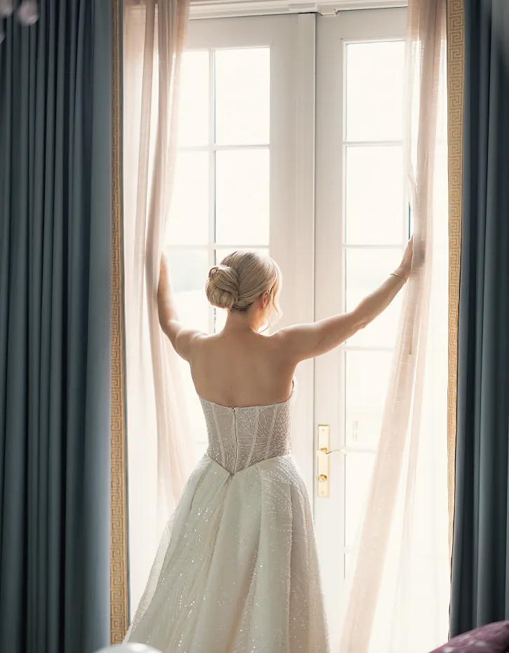 Bride opening sheer curtains at a window